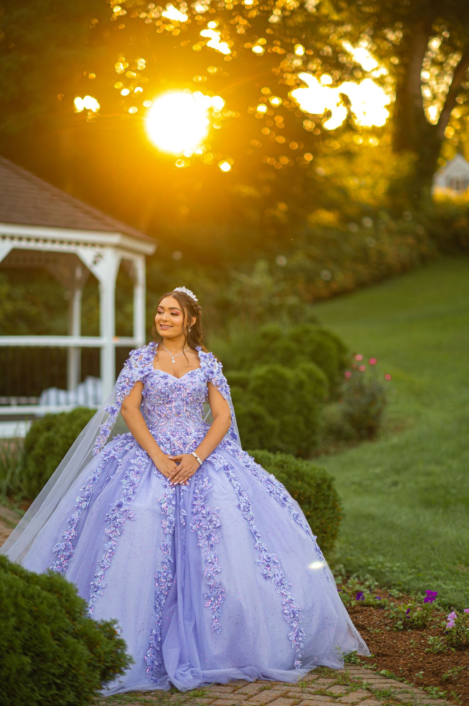 A lavender ball gown with floral appliques photographed at sunset near a white gazebo.