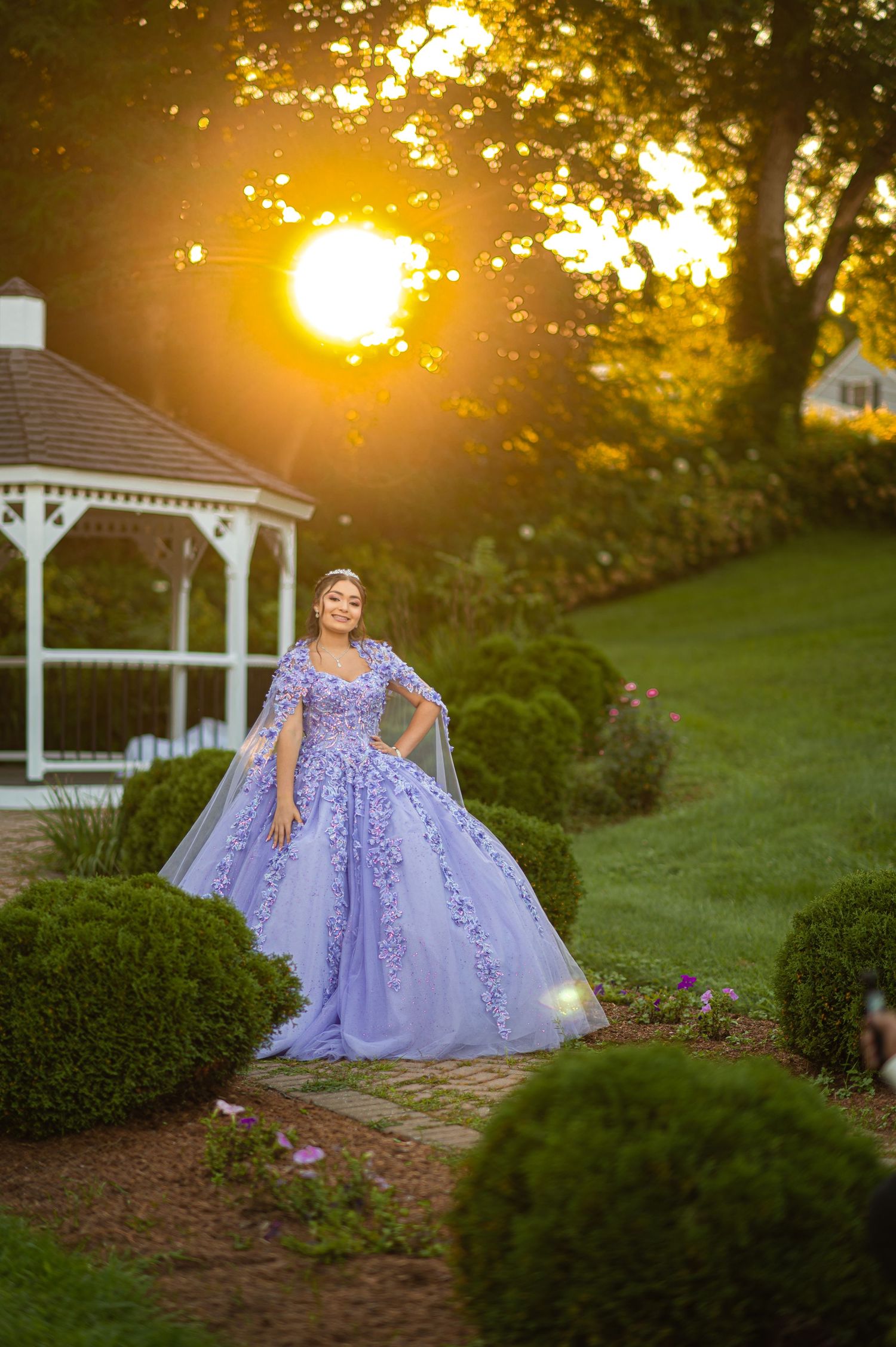 Elegant purple ball gown photographed in garden setting with gazebo and golden sunset behind.