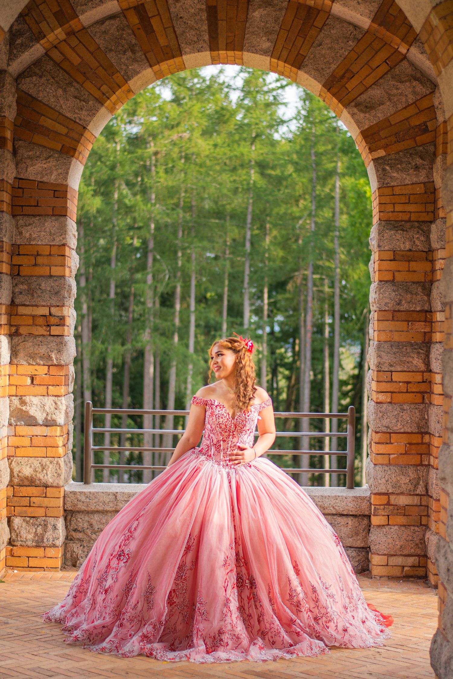 A pink princess ball gown photographed under a rustic brick archway with forest backdrop.