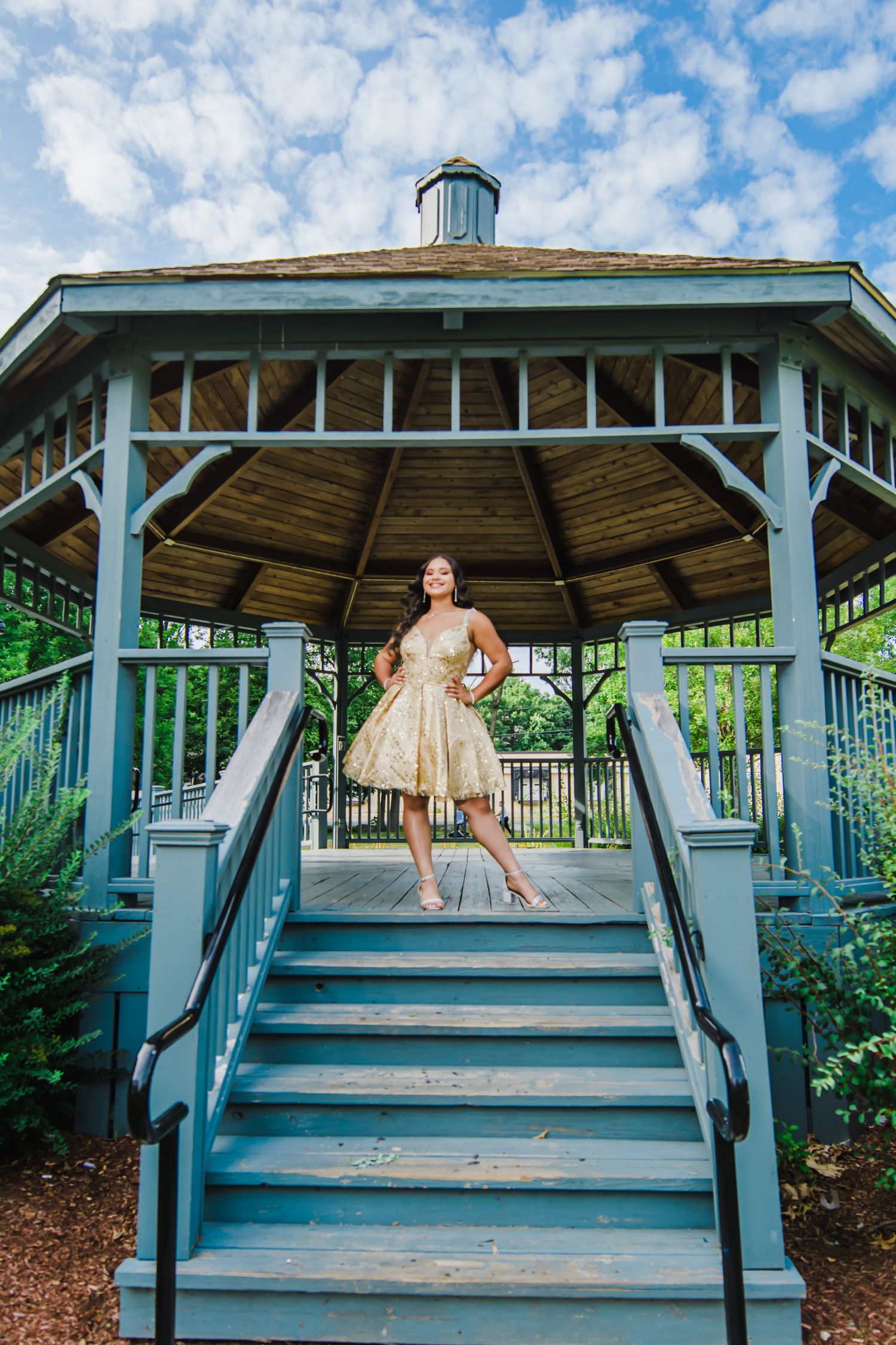 A short champagne-colored party dress shown in a white wooden gazebo with blue sky background.