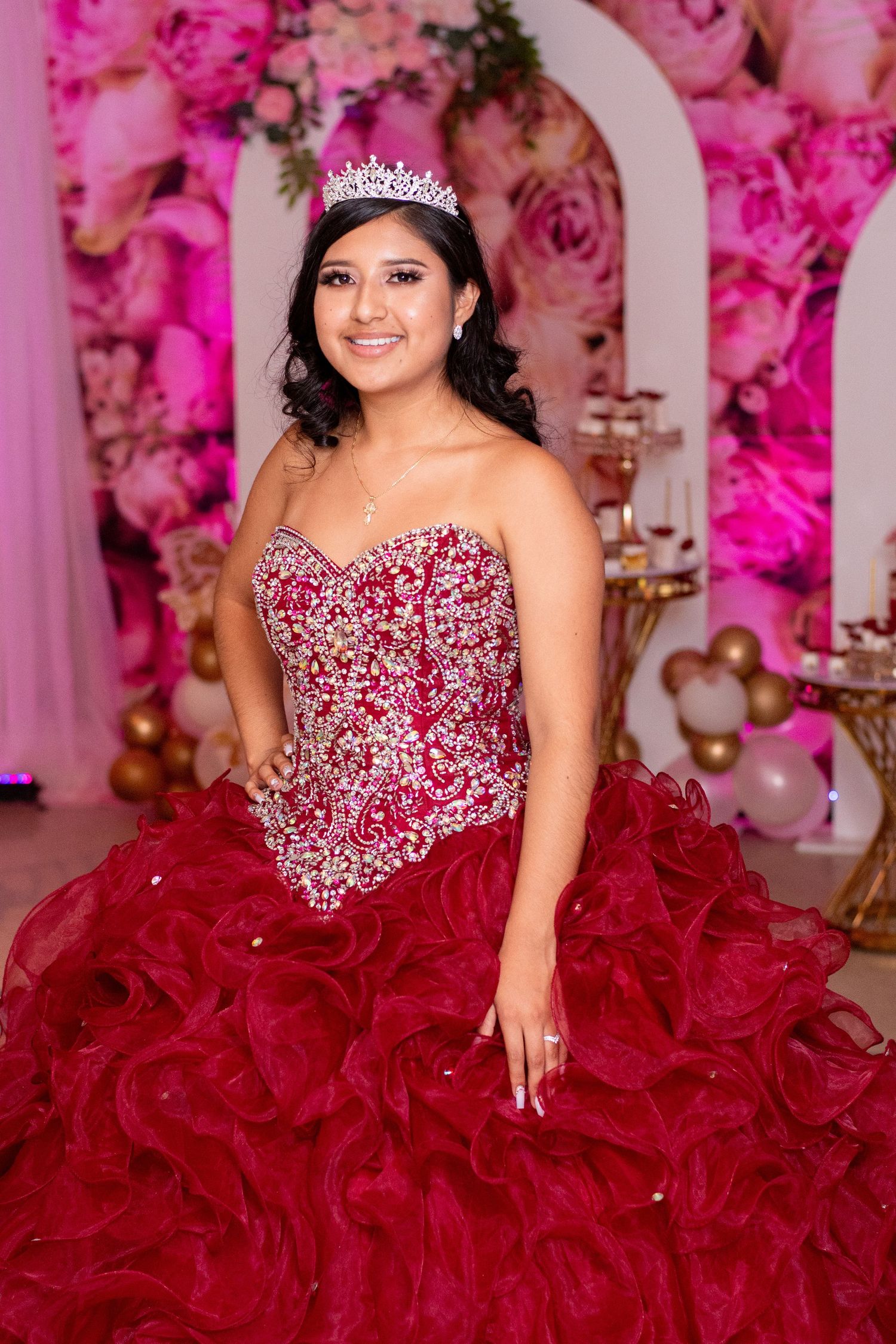 A red crystal-embellished ball gown photographed against a pink floral backdrop with balloons.