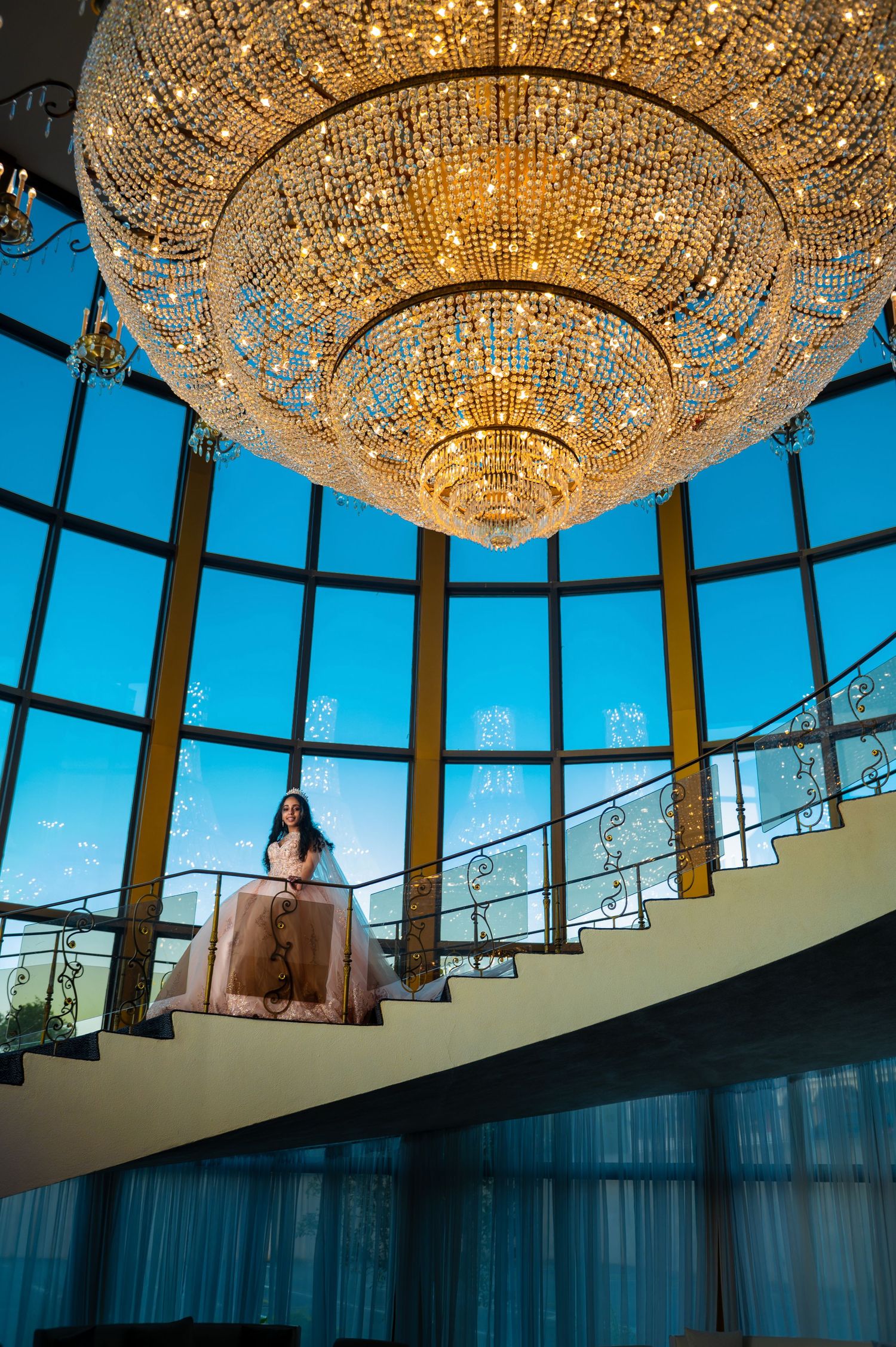 Grand crystal chandelier illuminates elegant staircase in modern venue with floor-to-ceiling windows.