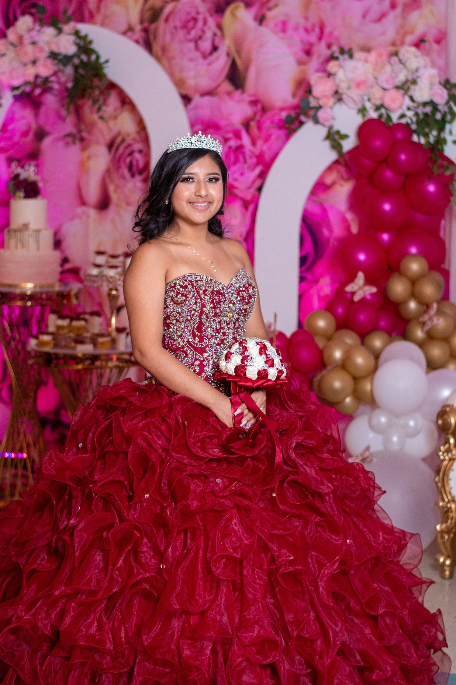A burgundy ball gown with crystal bodice photographed against a pink and gold balloon backdrop.