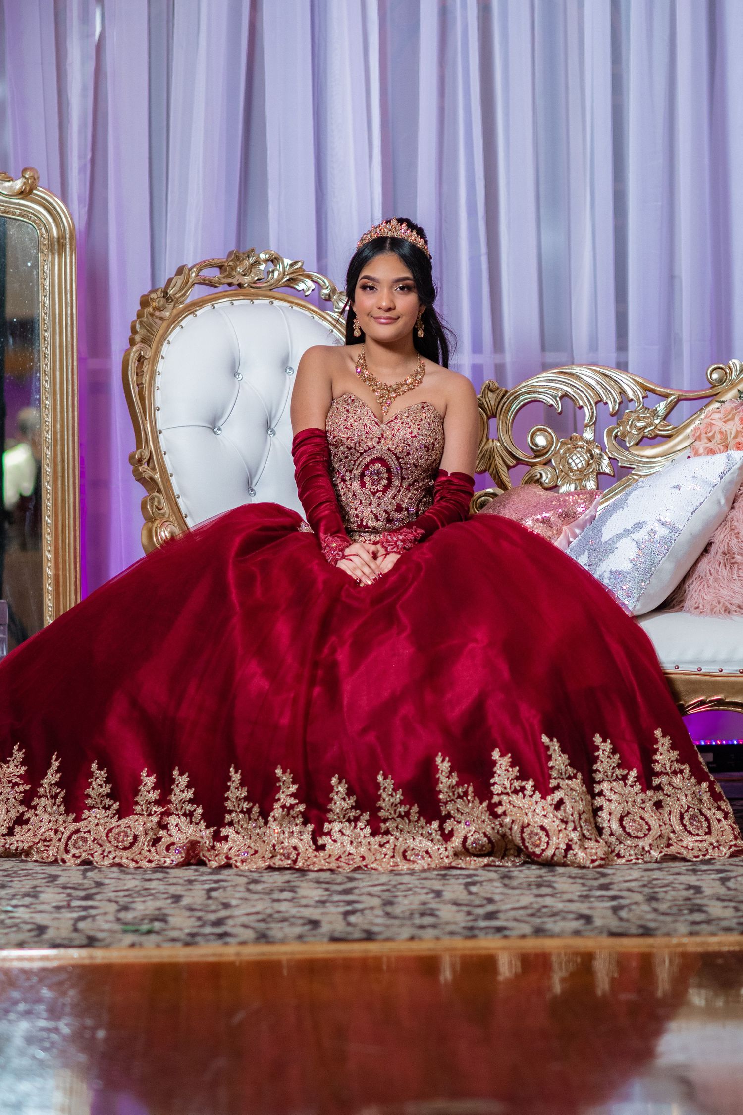 Luxurious red and gold quinceañera gown photographed on ornate white and gold throne.