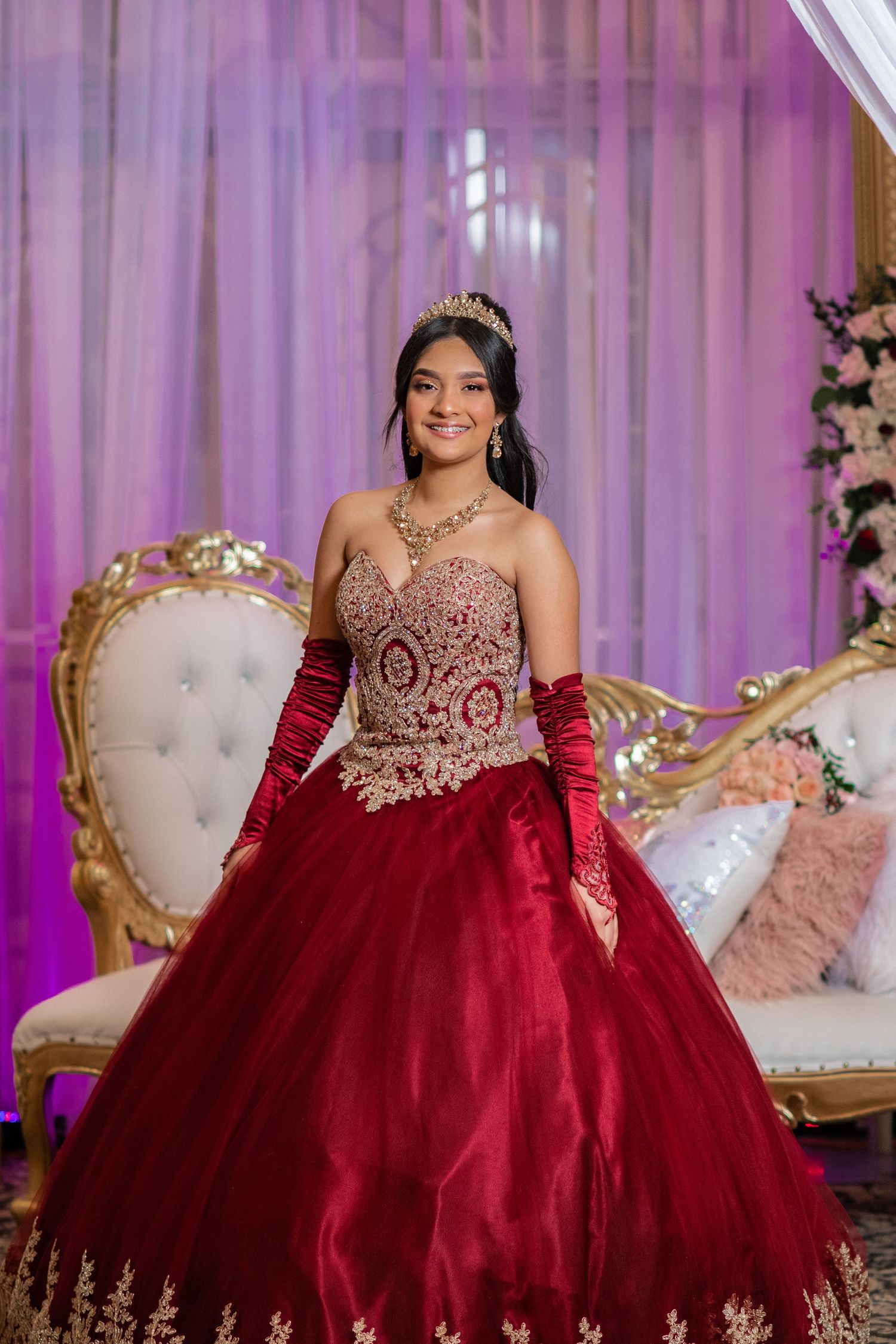 Stunning red ball gown with gold embellishments photographed against purple draped backdrop.