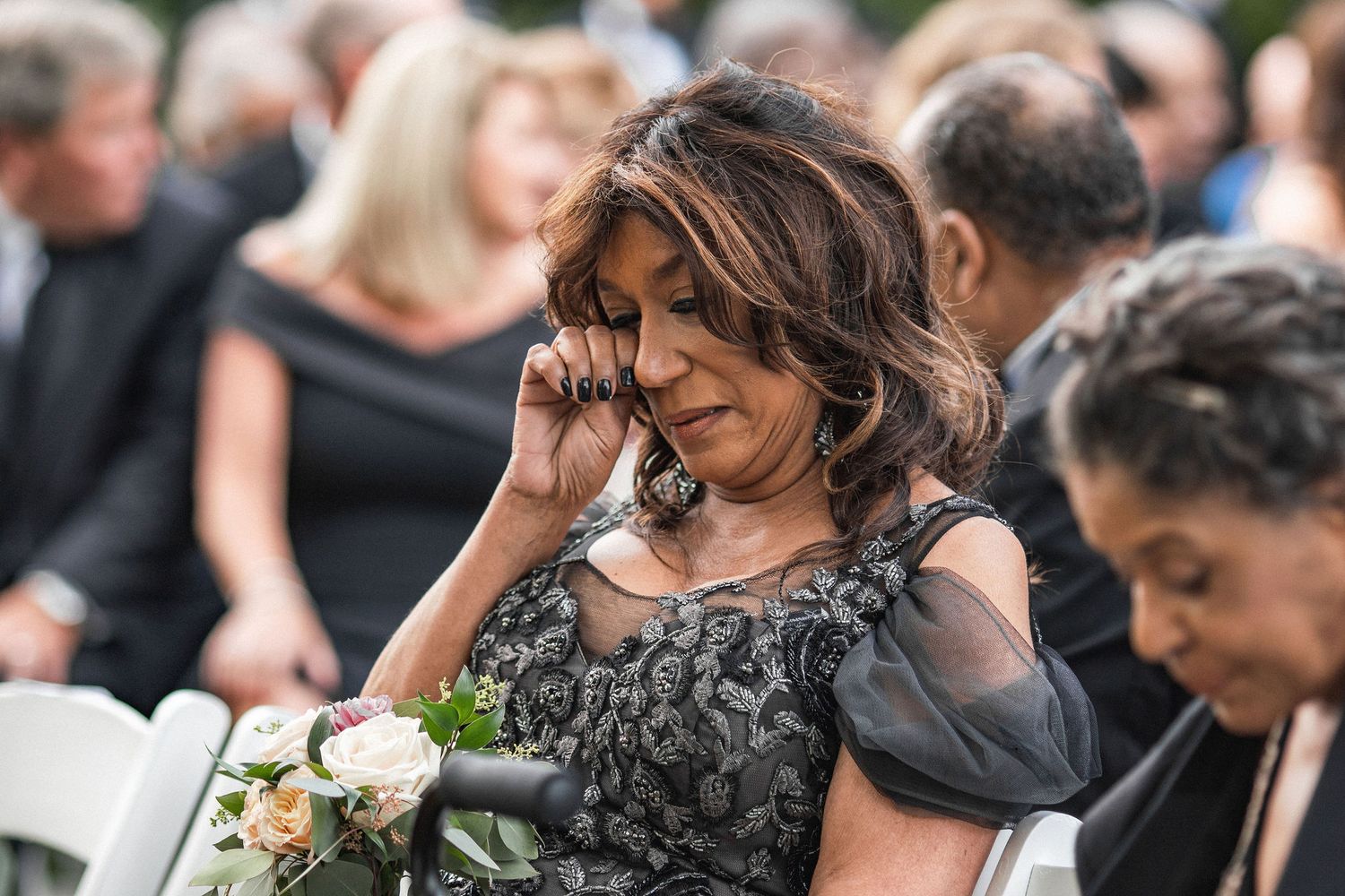 A wedding guest in a dark dress holding flowers while seated at an outdoor ceremony.