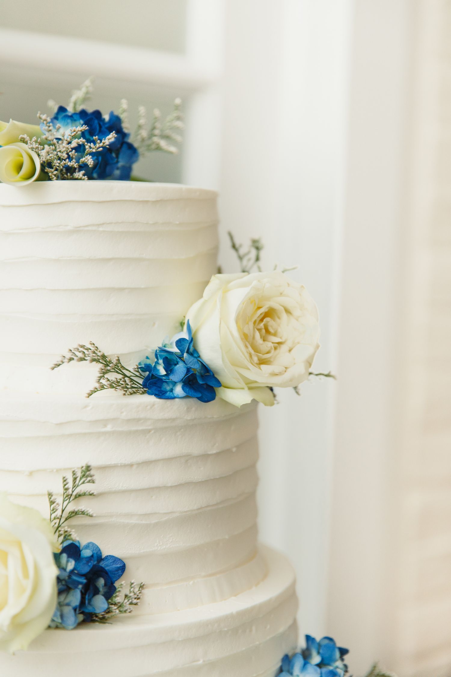 White wedding cake decorated with blue hydrangeas and cream roses against a light background.