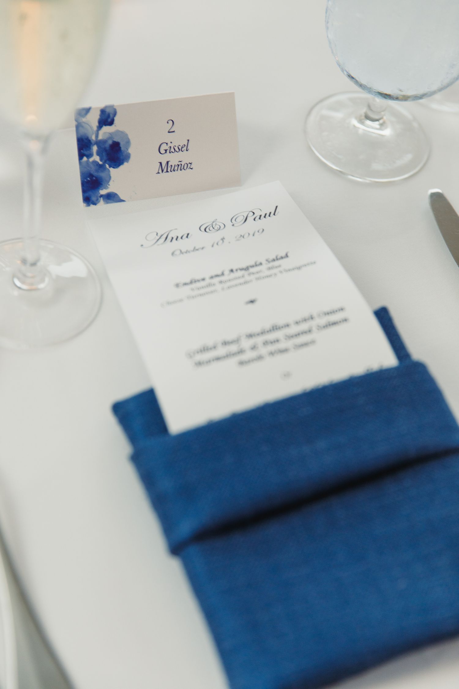 Wedding place cards with blue napkins and floral accents on a white tablecloth.
