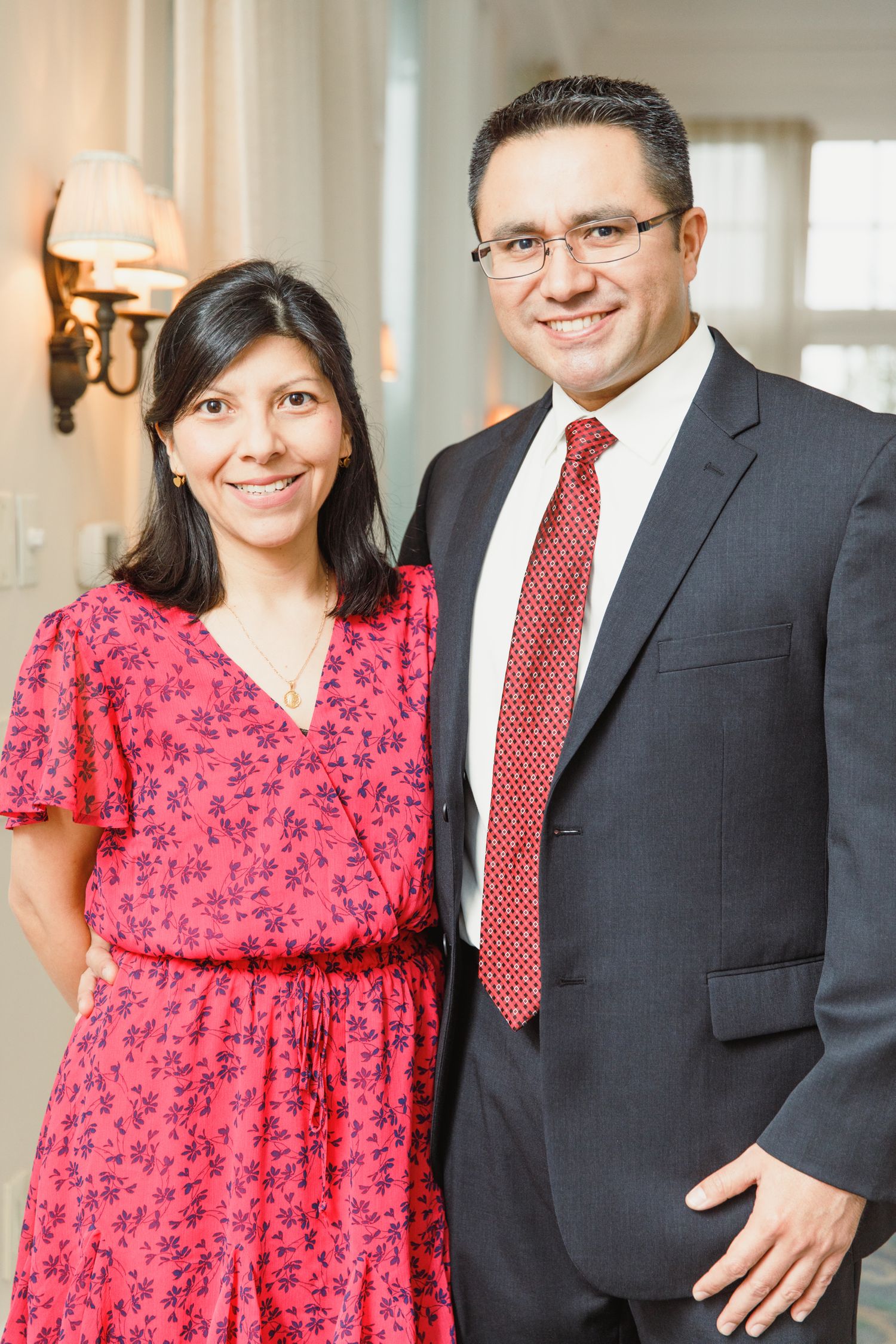 A couple in formal attire poses together in an elegant hallway with wall sconces.