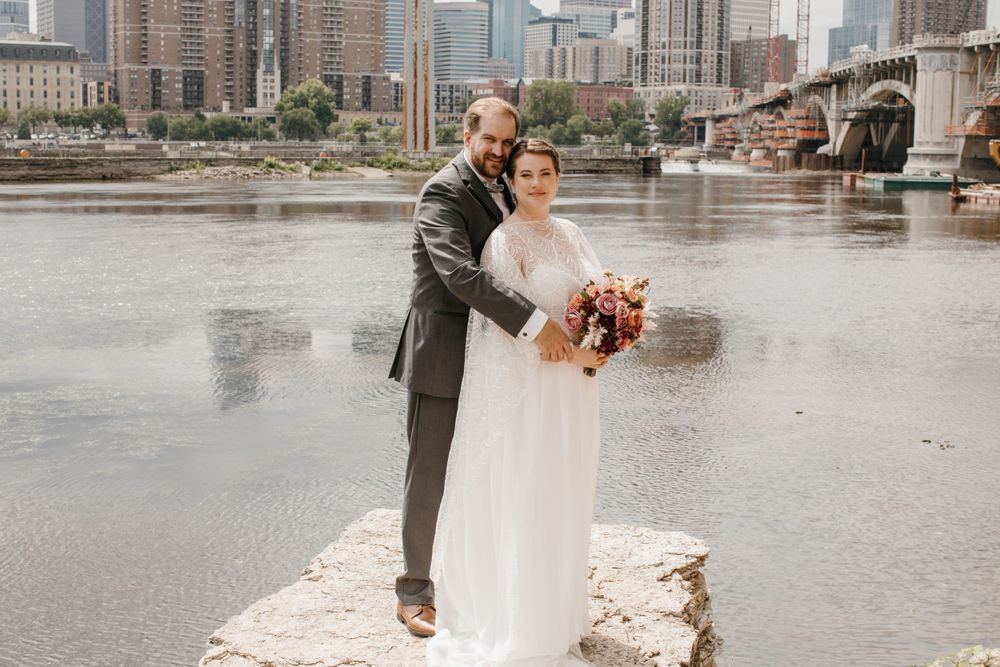 Bride and groom embrace on rocky riverbank with city skyline and bridge visible in background.