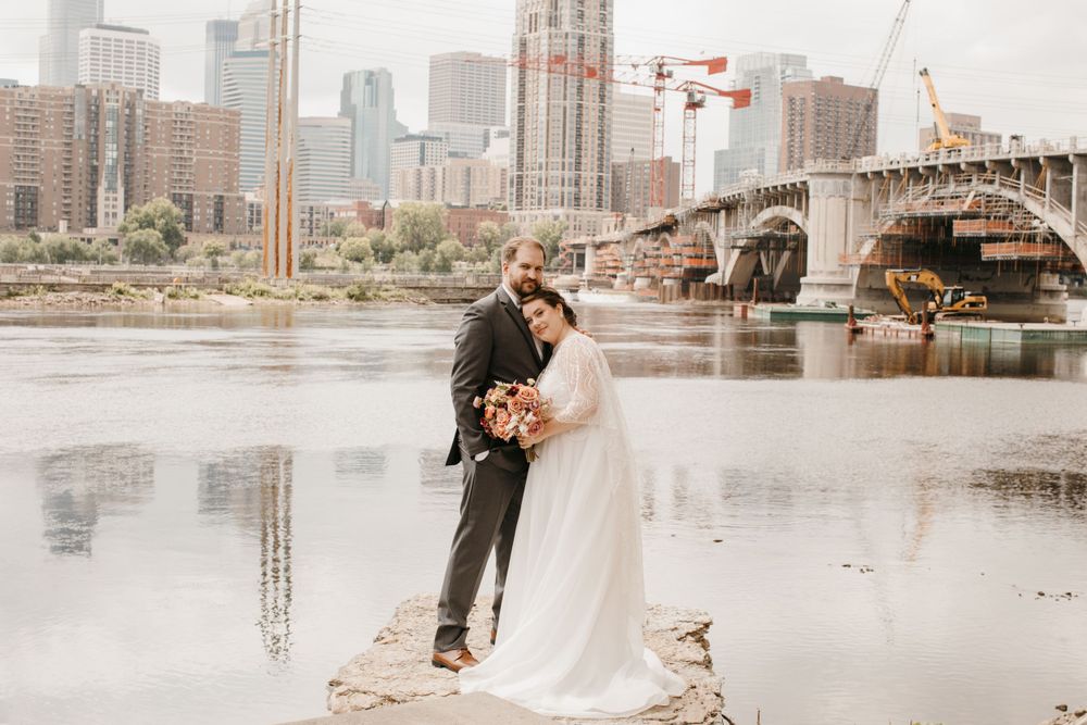 Groom embraces bride from behind on river rocks with urban skyline and bridge in background.