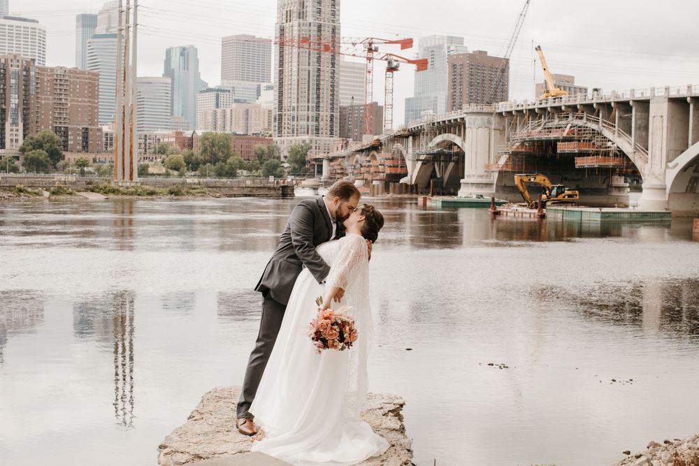 Groom dips bride for a kiss on rocky riverbank with city skyline and bridge reflected in water.