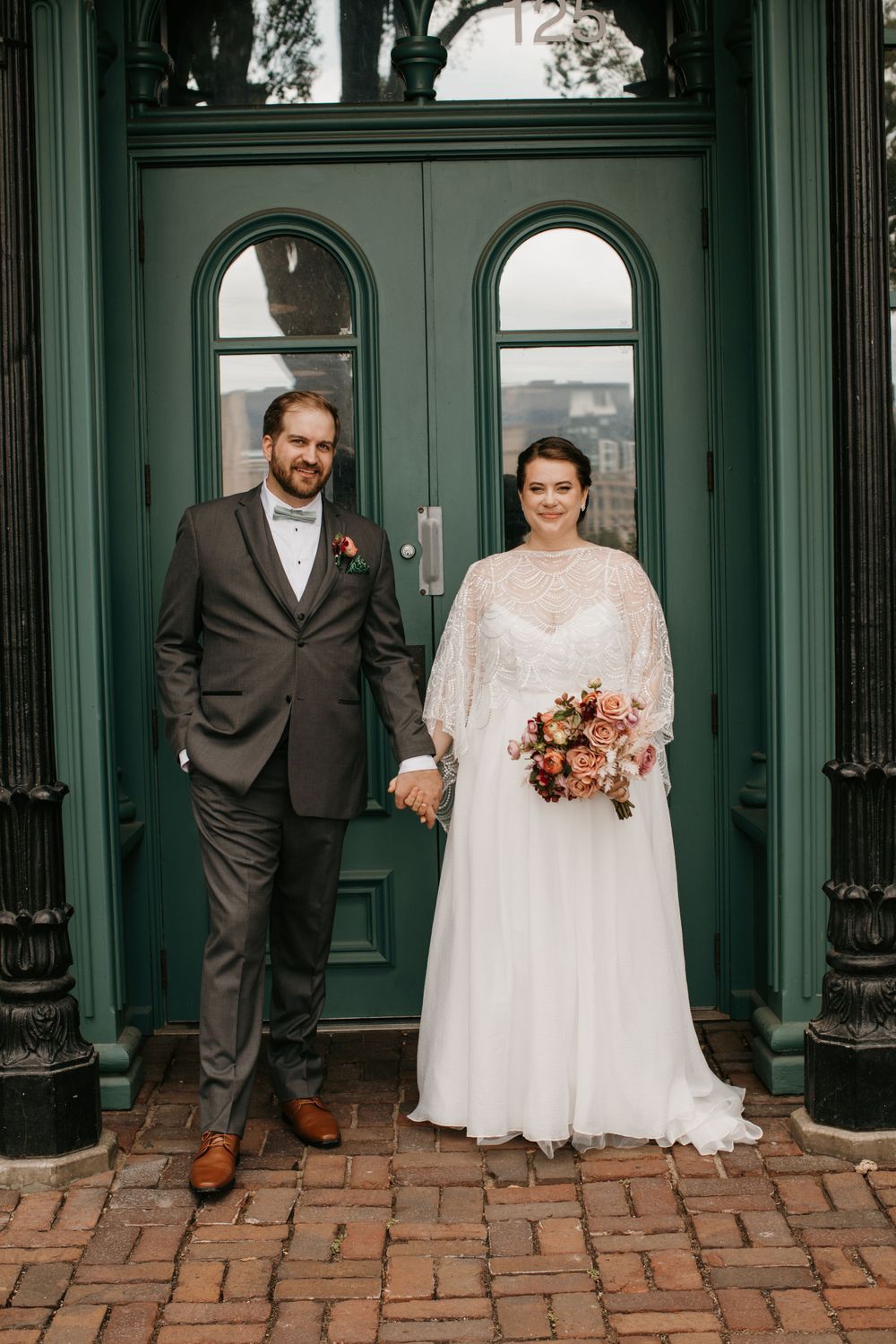 Newlyweds hold hands posing together in front of elegant green doors with arched windows.
