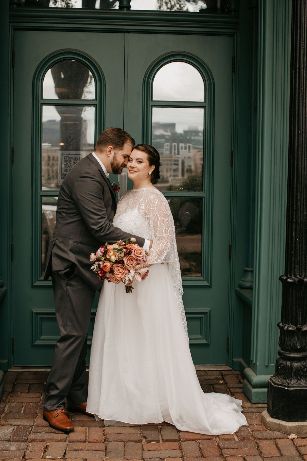 Groom tenderly embraces smiling bride holding autumn bouquet before green vintage doors.