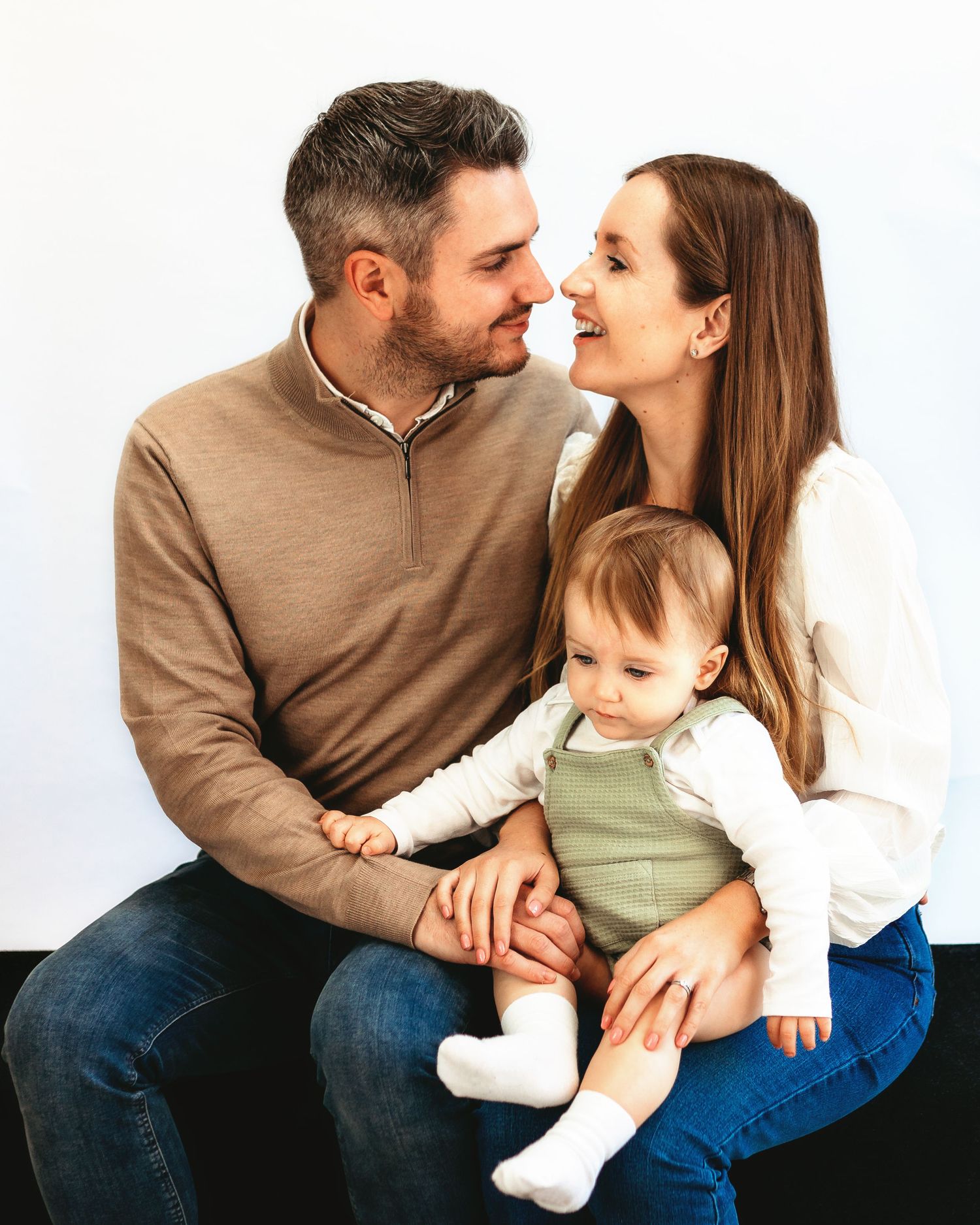 Family sitting close together on white backdrop sharing an intimate moment with their baby in olive green outfit.