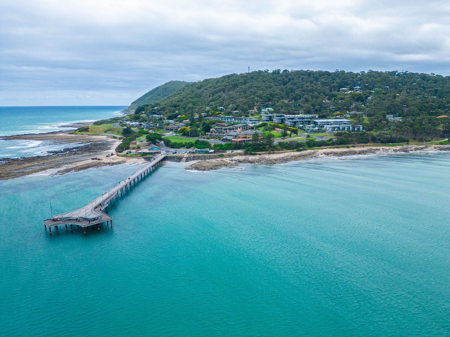 Aerial view of a wooden pier extending into turquoise waters with a green hillside and coastal homes in the background.
