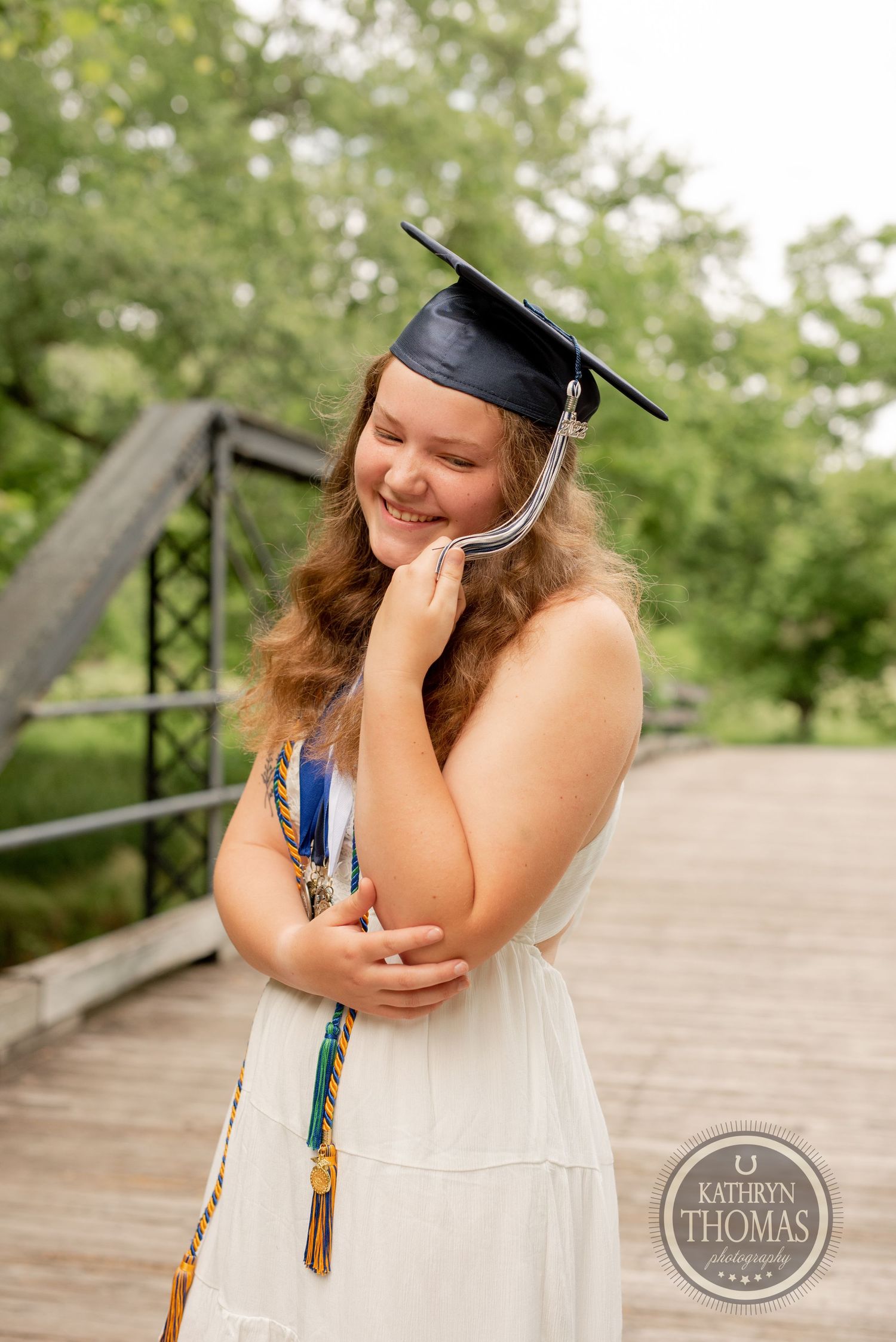 A graduate in cap and gown shows joy while posing on a wooden bridge.