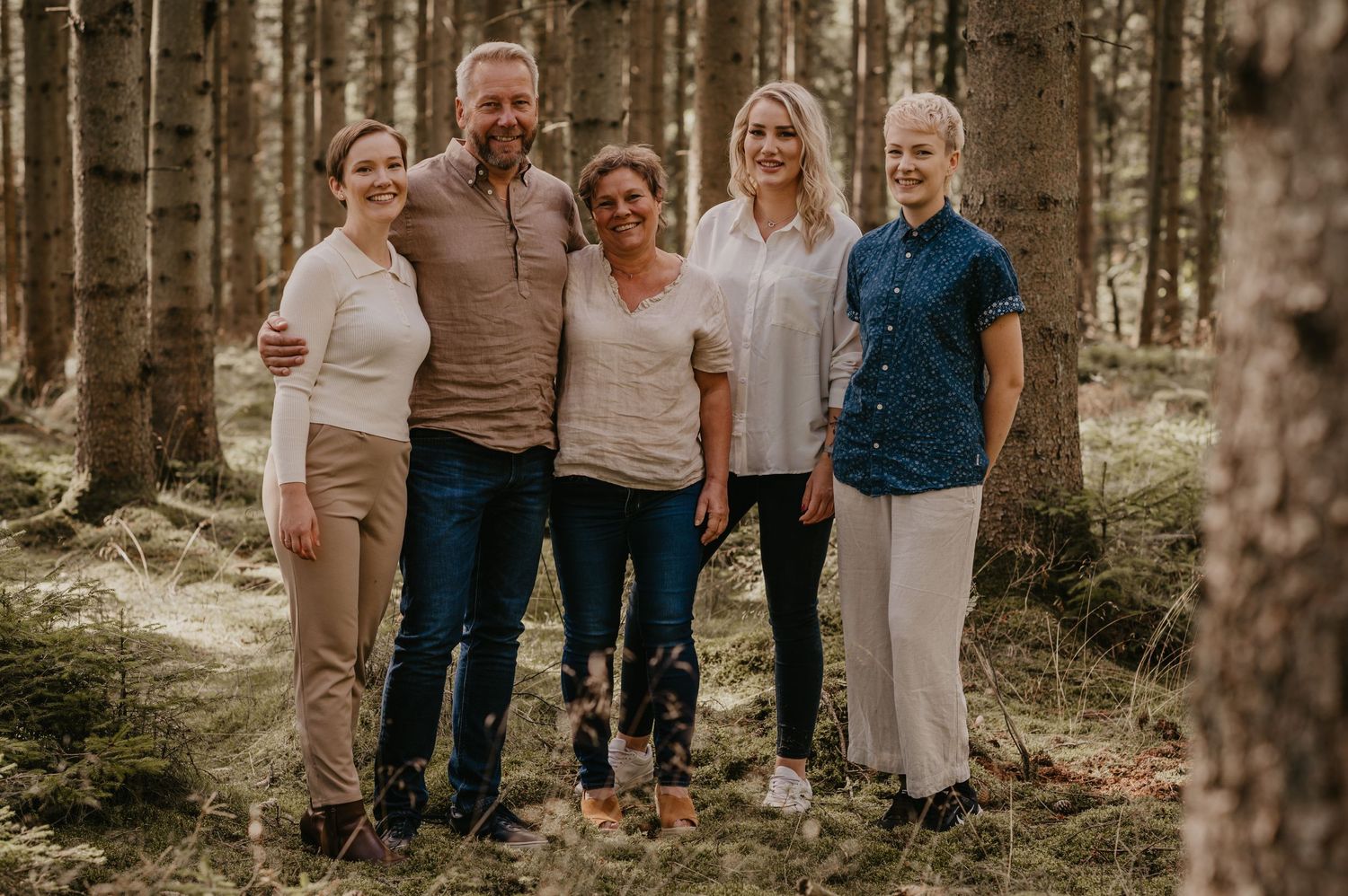 Five people smiling together in a sunlit pine forest, posing for a family portrait outdoors.