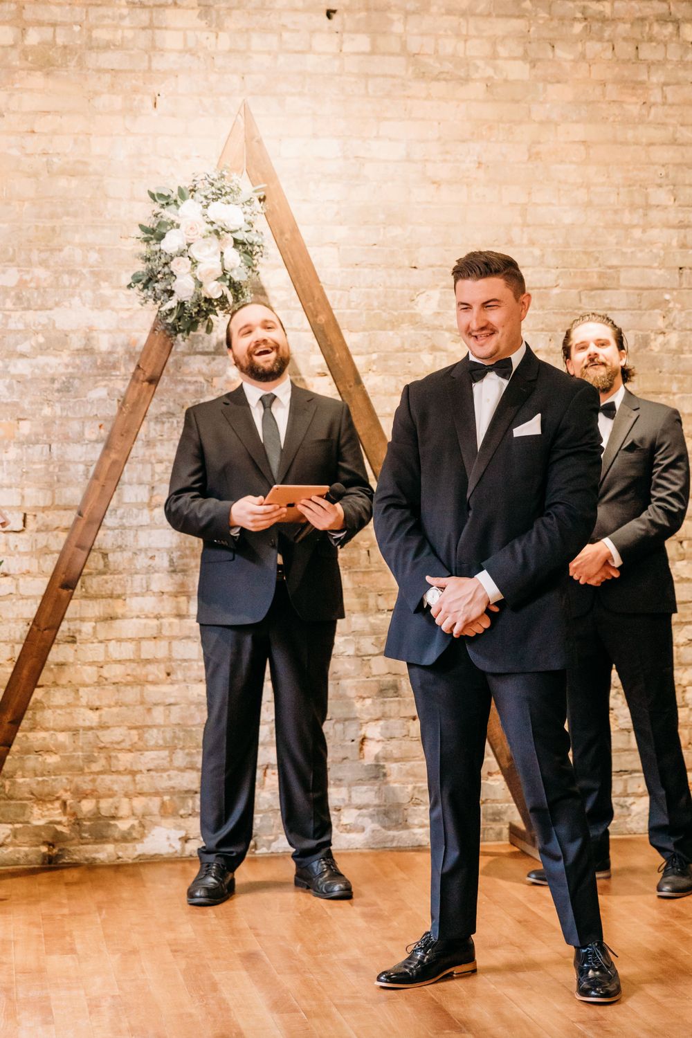 Wedding party members stand before a wooden triangle arch against exposed brick walls.