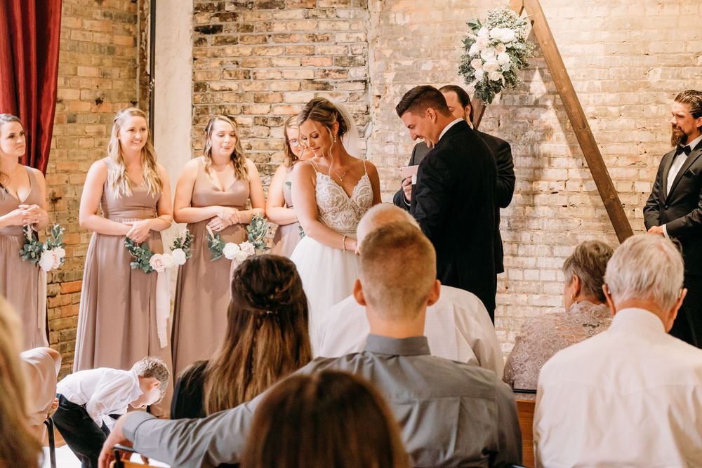 Wedding ceremony in progress with bridesmaids in mauve dresses against brick wall backdrop.