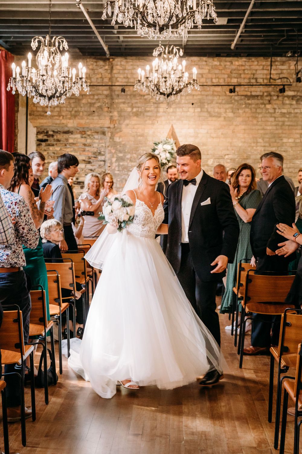 Newlyweds walking down aisle in rustic venue with chandeliers as guests applaud.