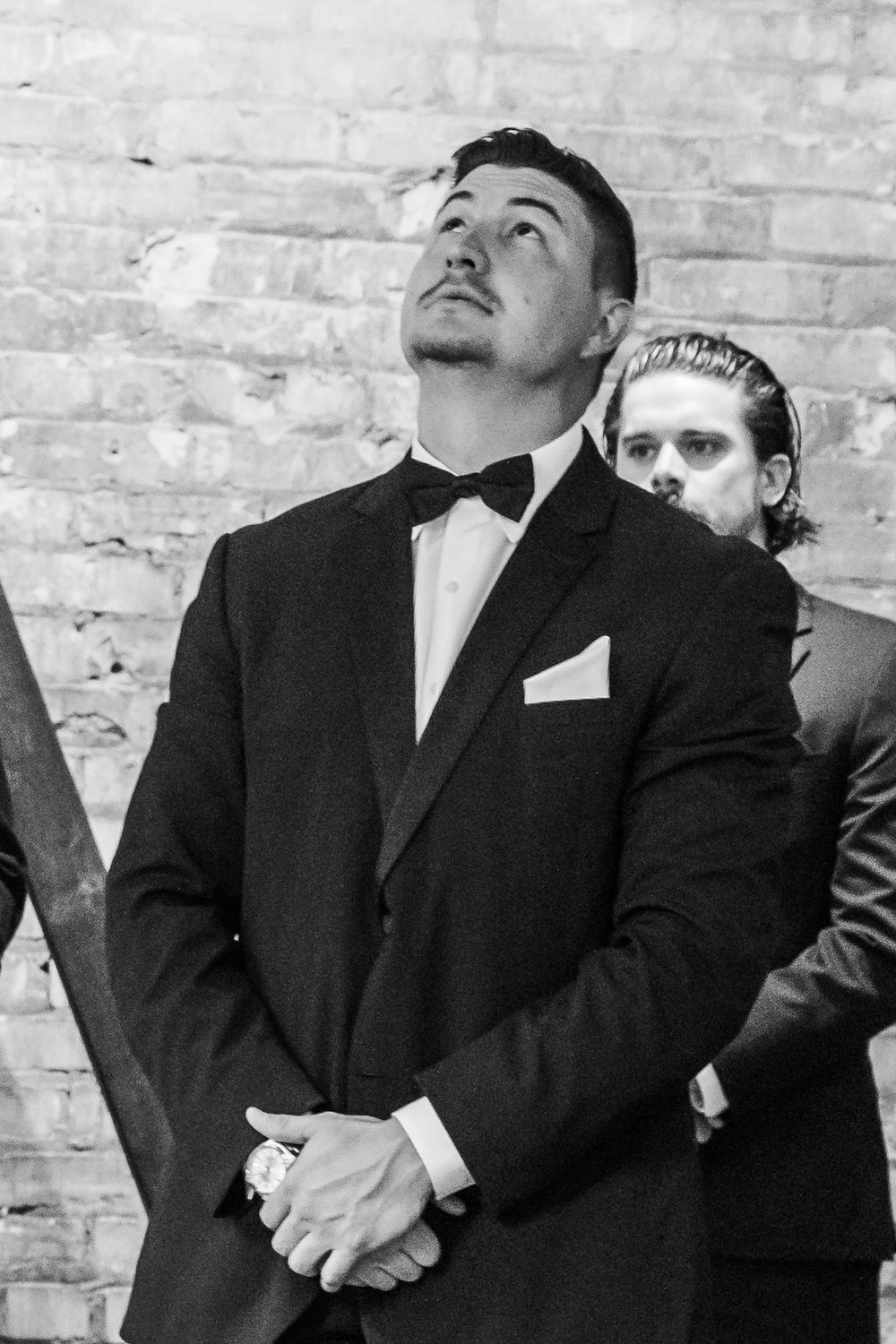 Groomsmen in black suits look upward with amused expressions against a brick wall backdrop.