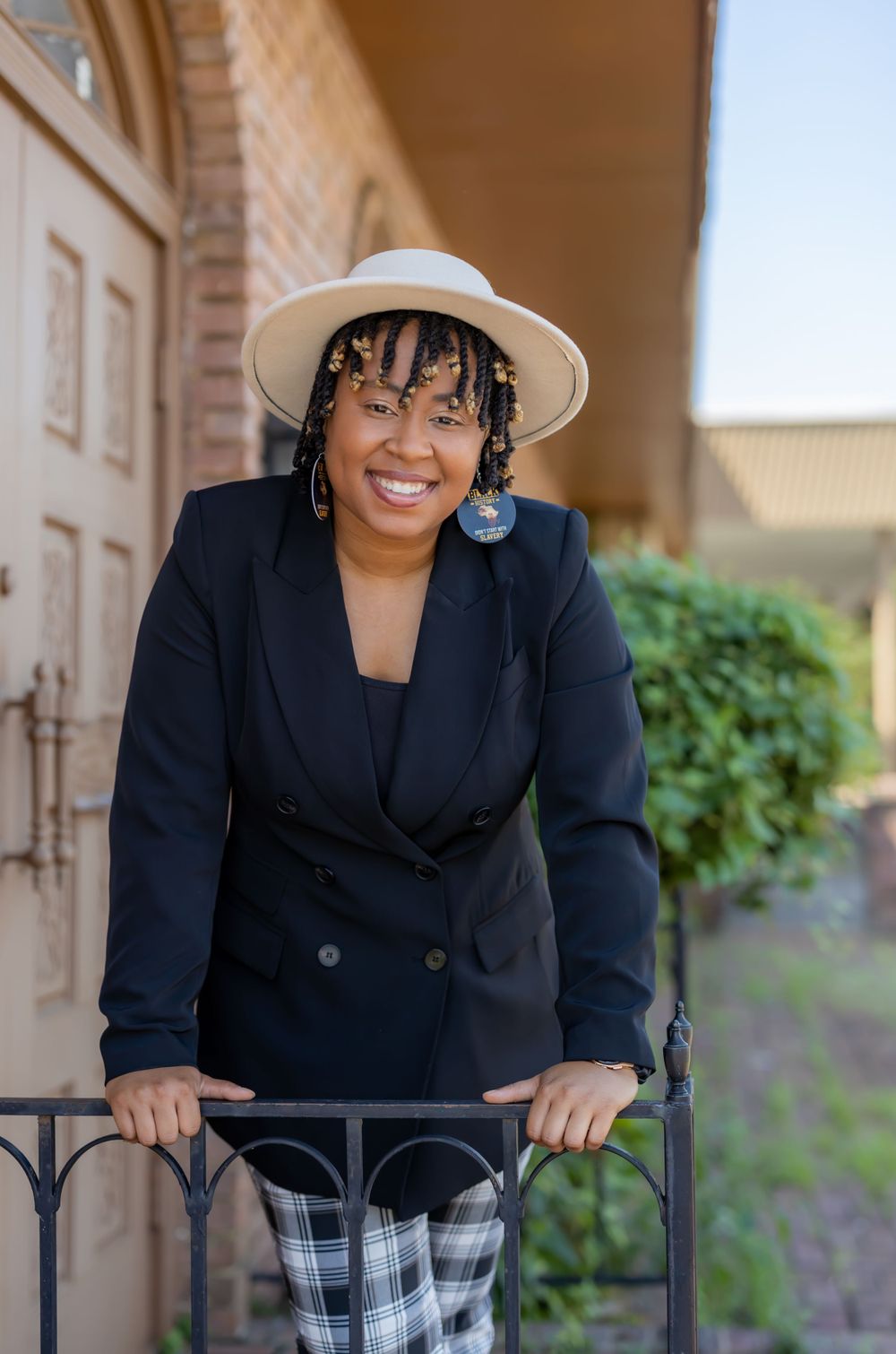 A stylish person in a black blazer and white hat leans on an iron railing with a building facade background.