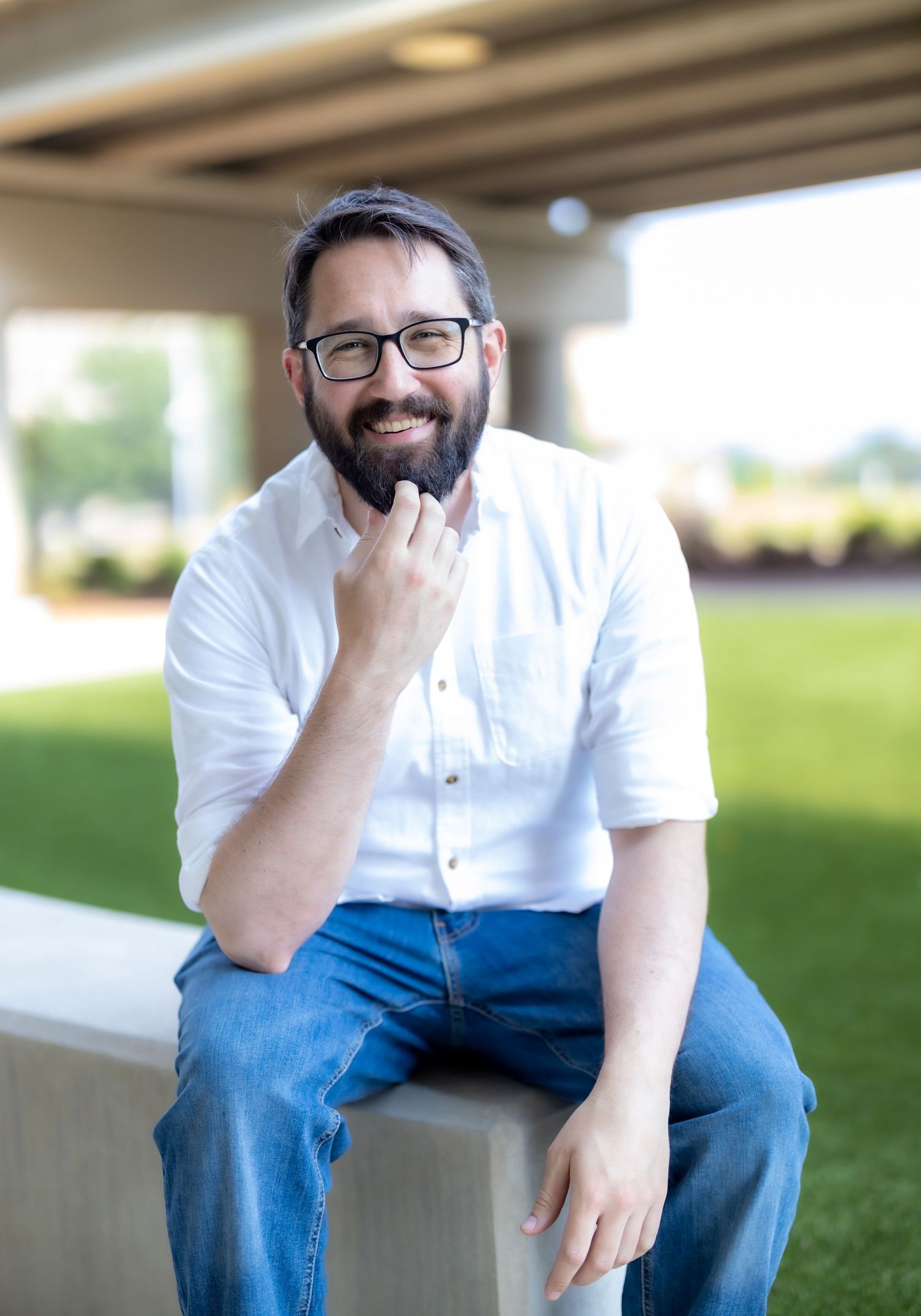 Person in casual attire with glasses sits thoughtfully on concrete ledge outdoors.