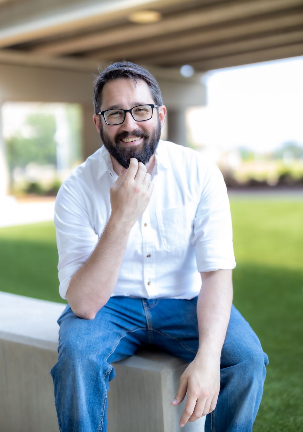 Person in casual attire with glasses sits thoughtfully on concrete ledge outdoors.