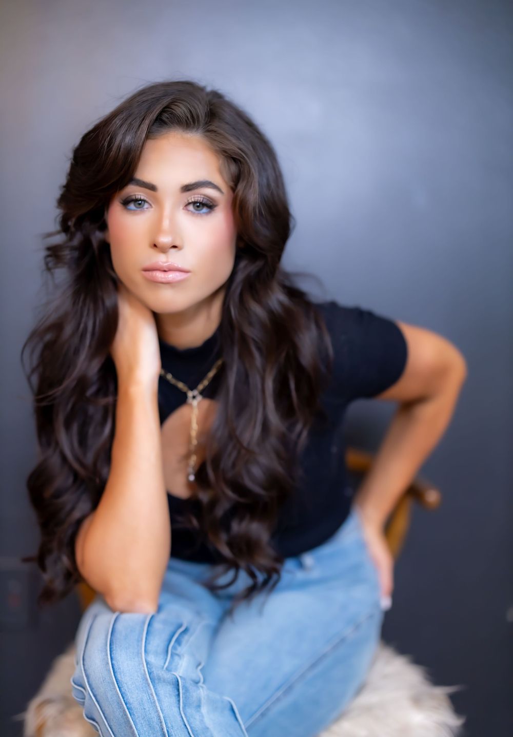 Dark-haired model in black shirt and jeans poses for dramatic studio portrait against grey background.