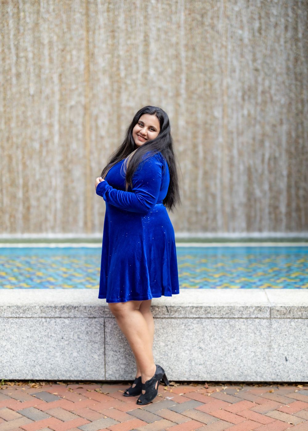 Model in royal blue velvet dress poses elegantly against textured wall backdrop.