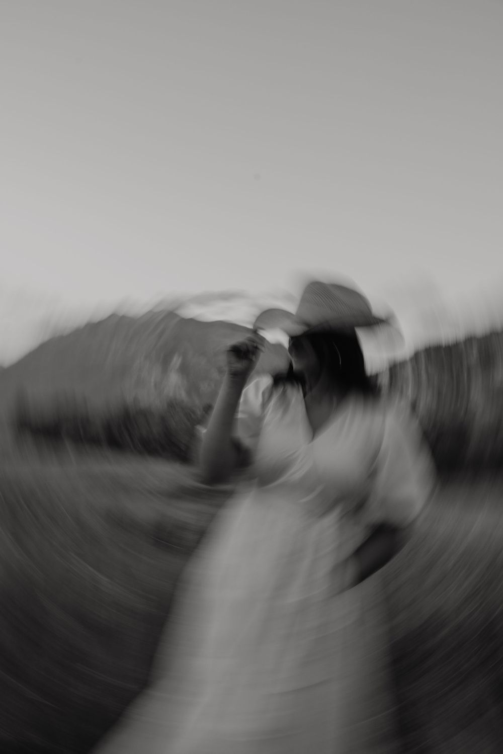 Artistic black and white motion blur photo of a figure in white dress and hat in an outdoor setting.