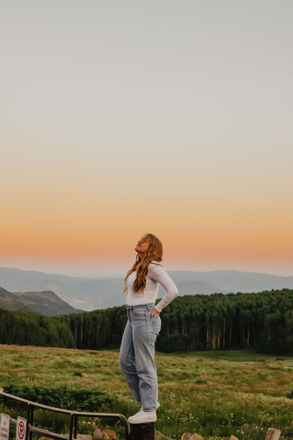 Figure in casual wear stands on railing overlooking mountain valley at sunset.