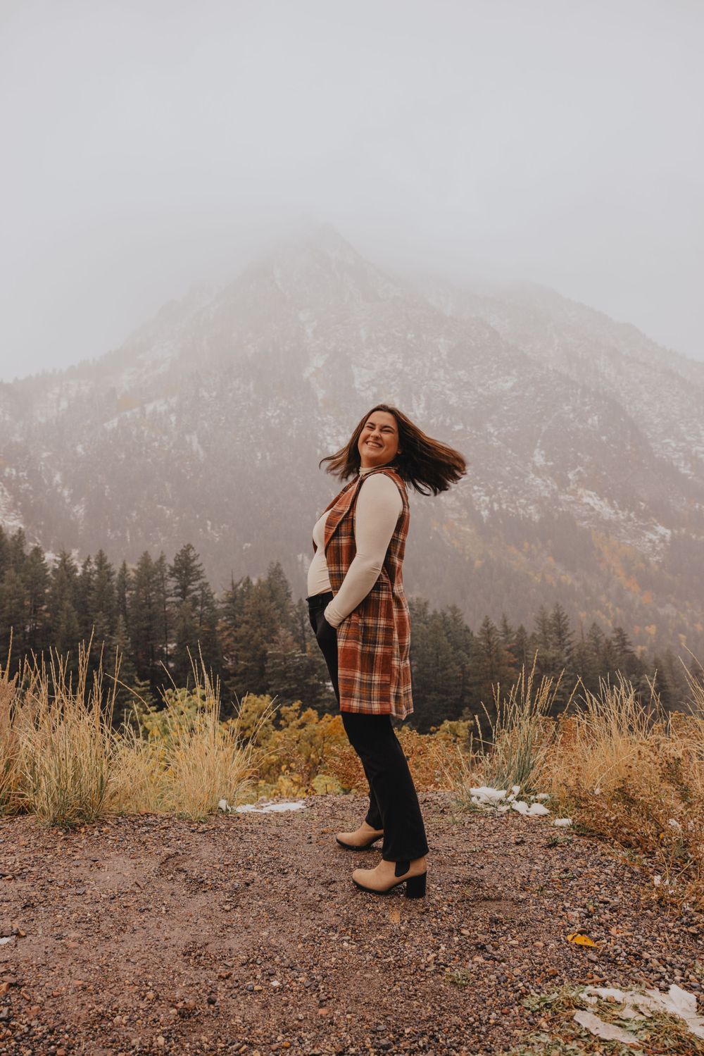 Woman in plaid vest twirling on mountain trail with autumn foliage and snowy peaks.