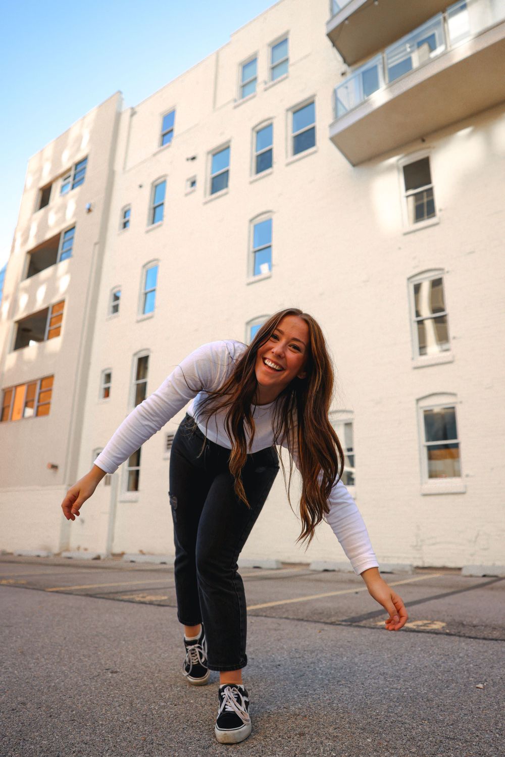 Playful pose in white long sleeve and dark pants against beige apartment building.
