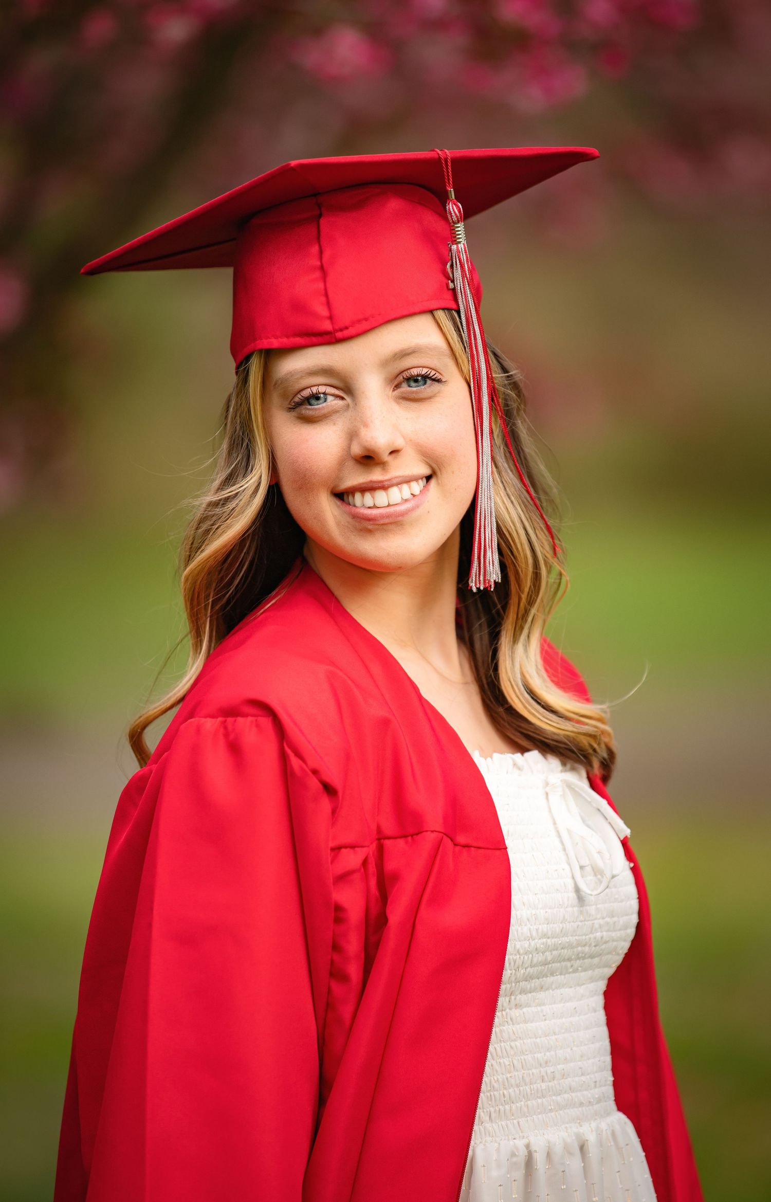 Graduate in red cap and gown smiling outdoors with cherry blossoms in background.