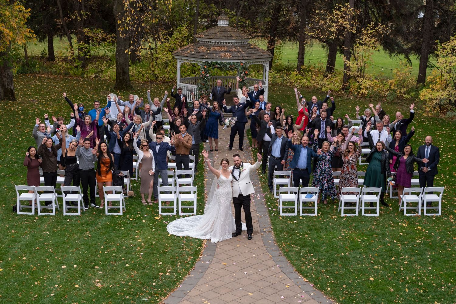 Aerial view of outdoor wedding ceremony under wooden gazebo with seated guests on white chairs surrounded by autumn trees.