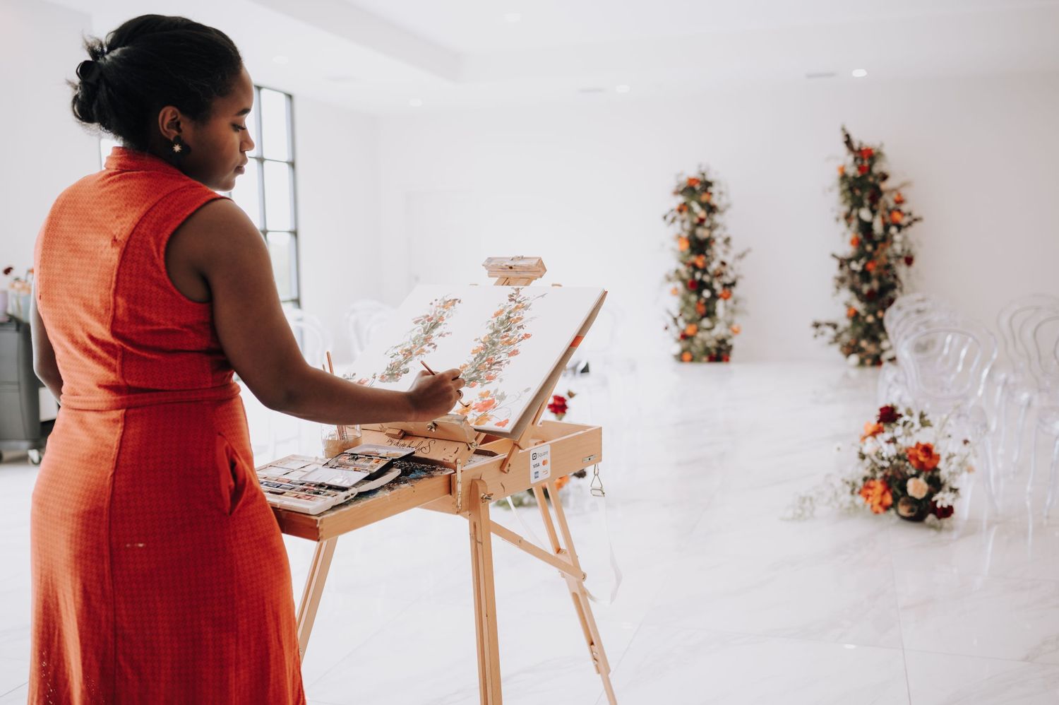 Artist in red dress painting at easel with floral arrangements in background.