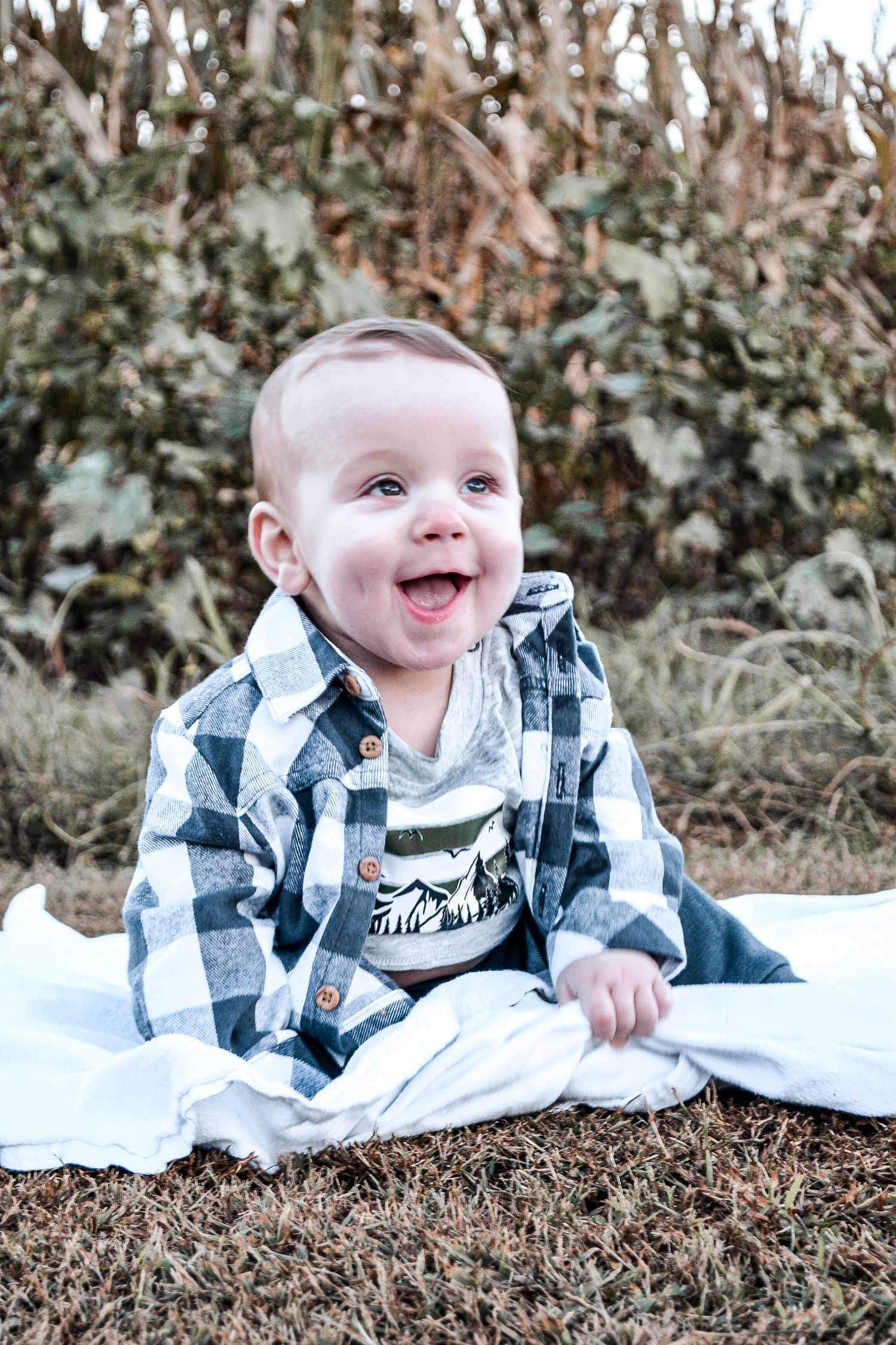 A smiling baby in a plaid shirt sits on a blanket outdoors against green foliage.