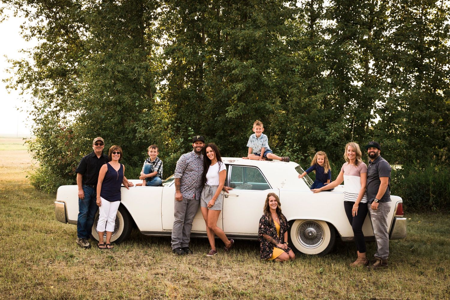 Large family group posing around a vintage white car in a sunny field.