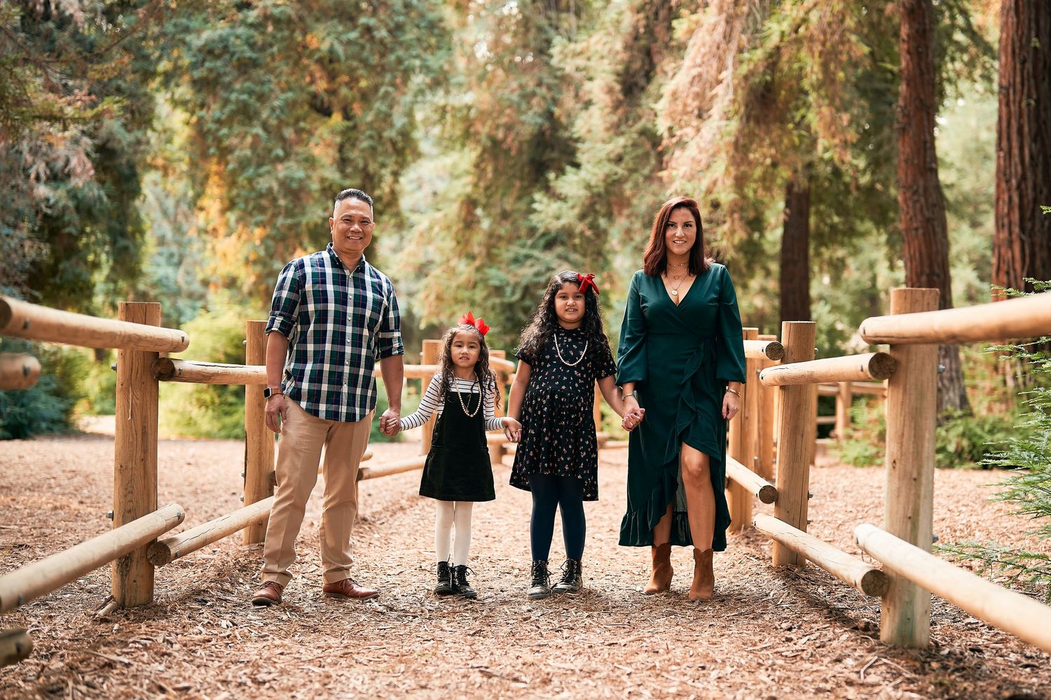 Family of four standing together on wooden bridge path surrounded by forest.