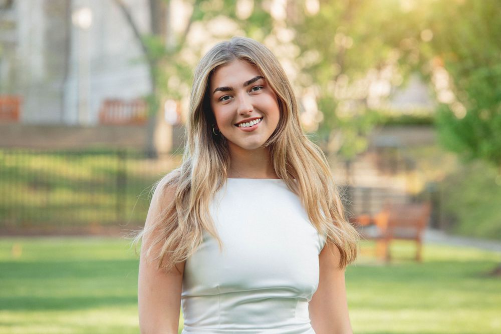 Woman in white dress smiles outdoors with sunlit campus greenery in background.