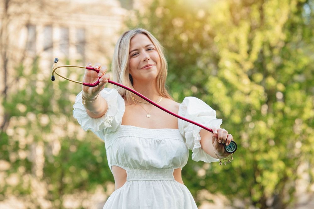 A woman in a white dress poses outdoors with a violin against a sunlit natural background.