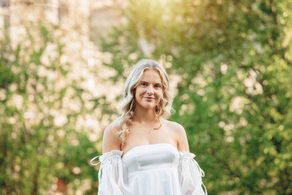 Woman wearing an elegant white off-shoulder dress poses in a garden setting with warm lighting.
