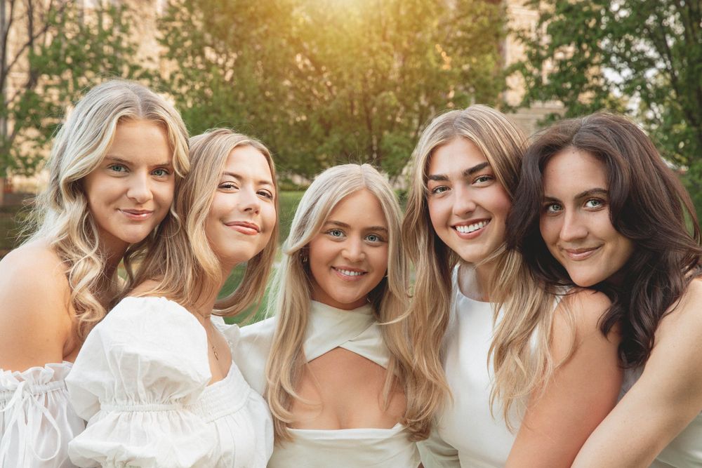 Group of friends in white dresses smile together during a sunny outdoor photoshoot.