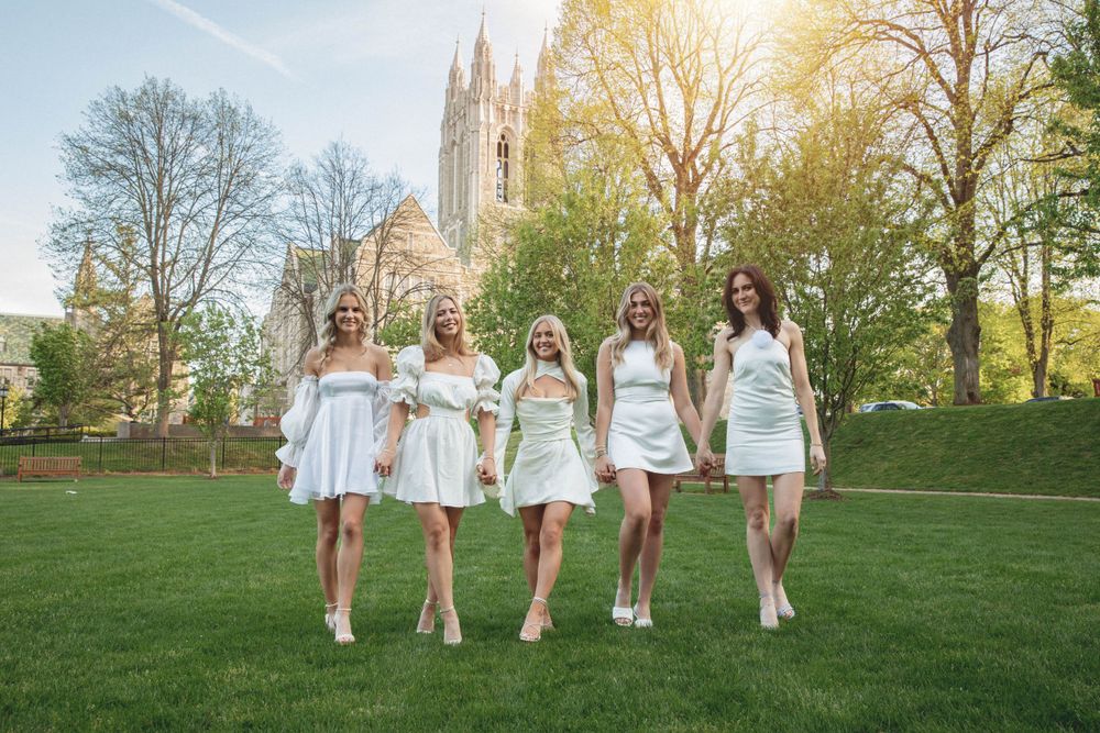 Five women in matching white dresses walk across lawn with Gothic church tower in background.