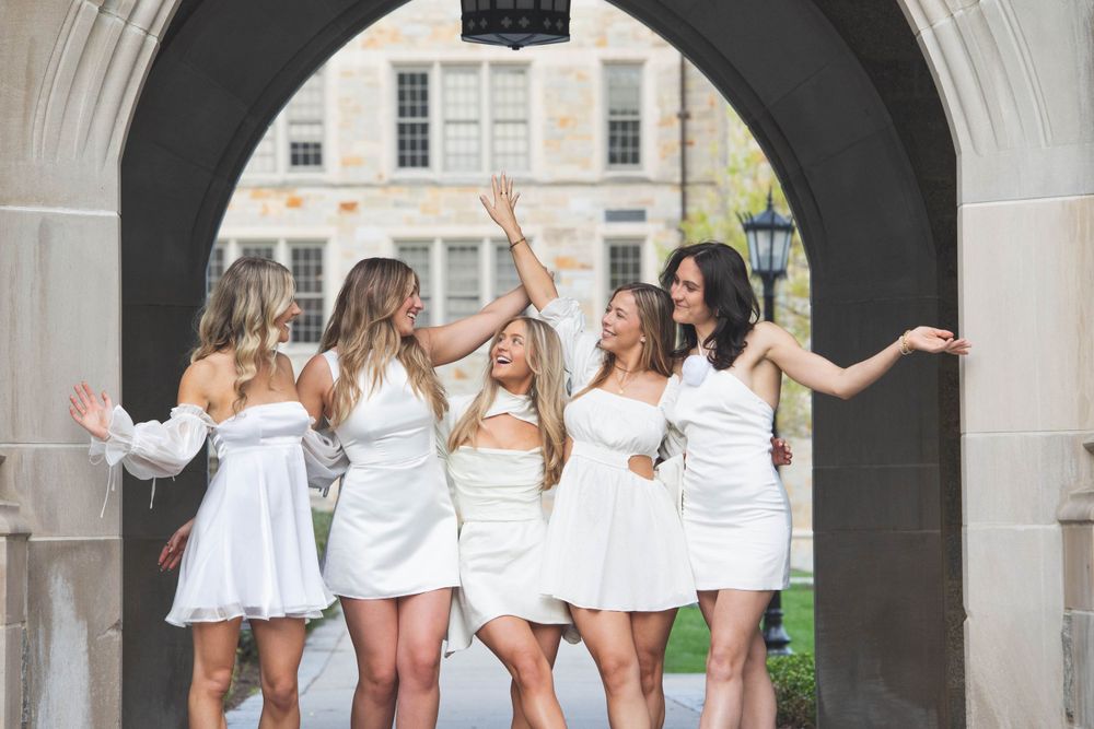 Friends in white dresses celebrating joyfully under an ornate stone archway.