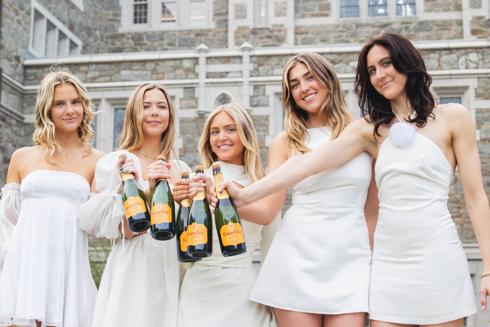 Five friends in white dresses toasting with champagne bottles on stone steps of a historic building.