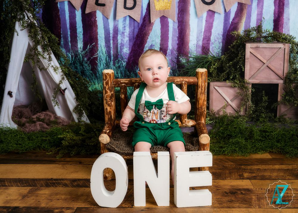 One year old in green outfit sitting on wooden chair with 'ONE' letters and forest backdrop.