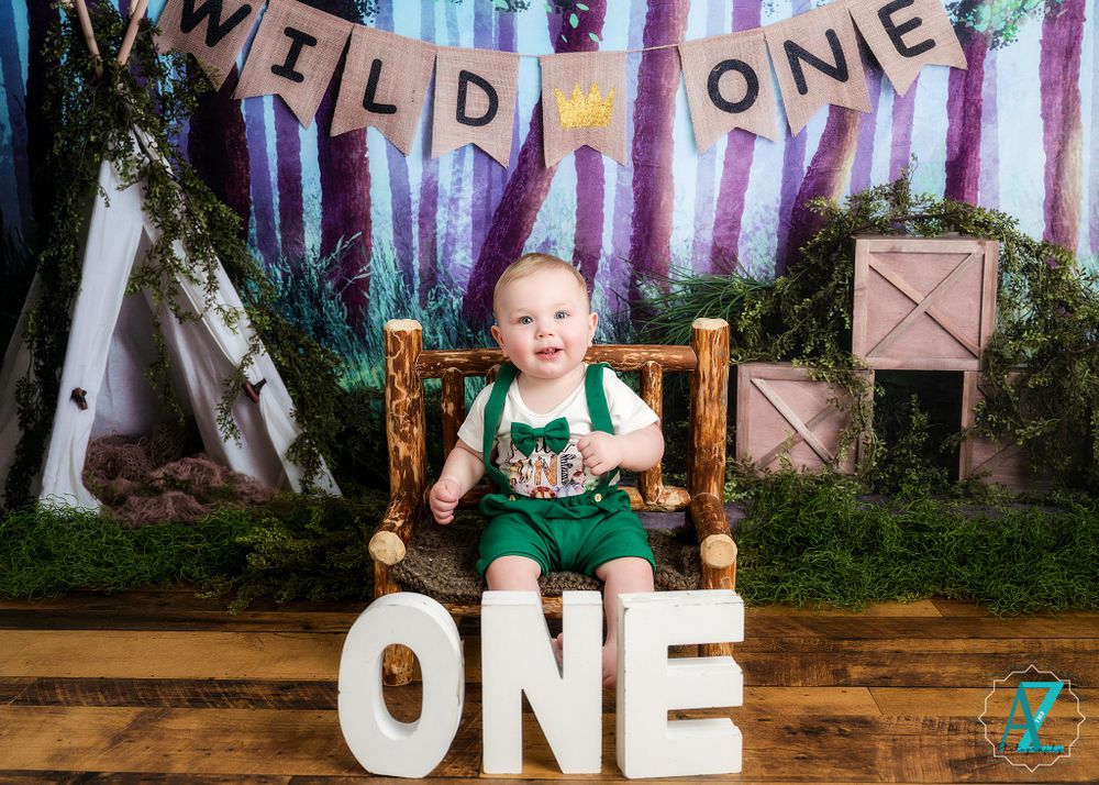 Smiling toddler in green outfit posed on rustic chair with 'Wild One' themed backdrop.